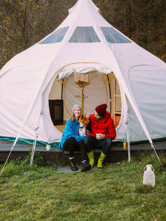 Couple-of-vacation-goers-campers-sit-in-front-of-big-glamping-tent-with-their-dog