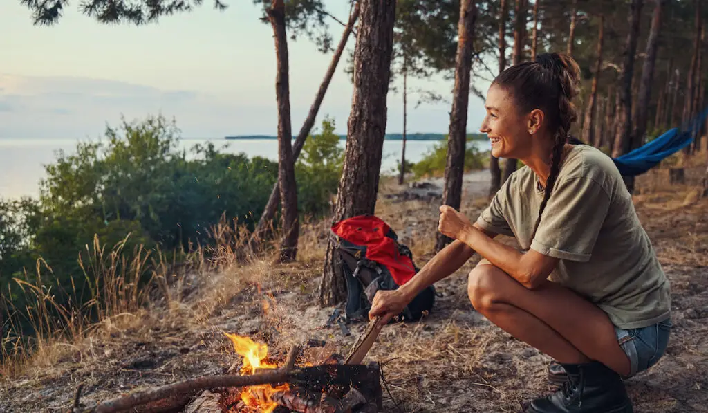 Happy traveler is adding firewood to campfire, camping concept