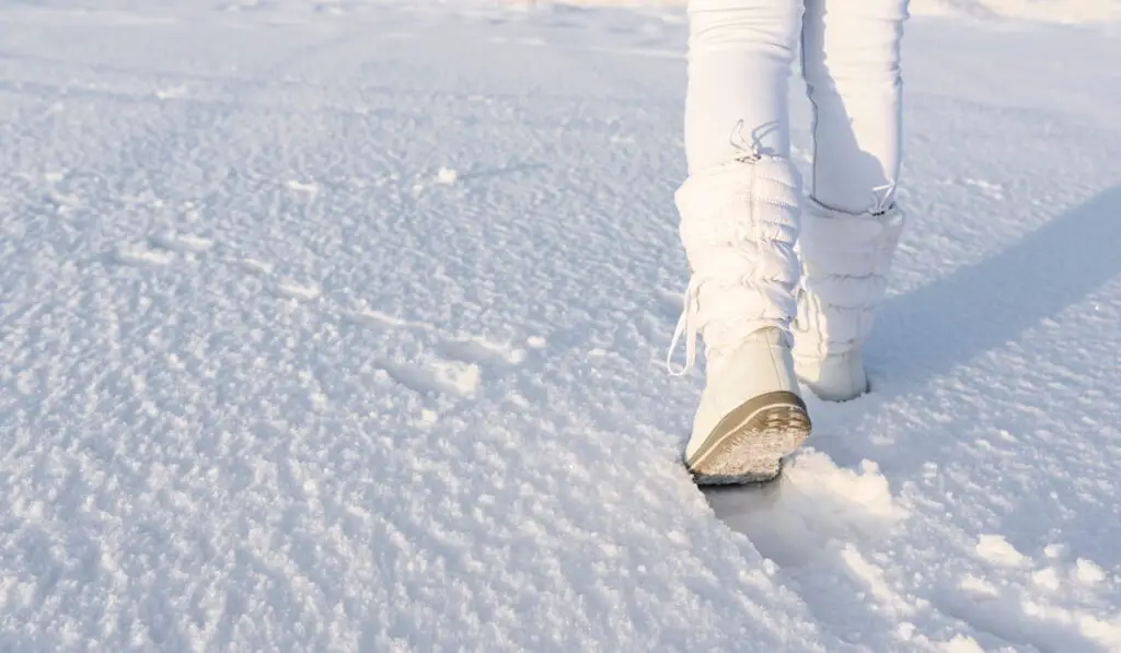 Low angle view of walking away female legs with white snow boots on, in deep snow. 