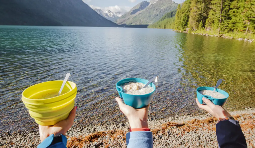 friends showing their oatmeal breakfast while camping near mountain lake 