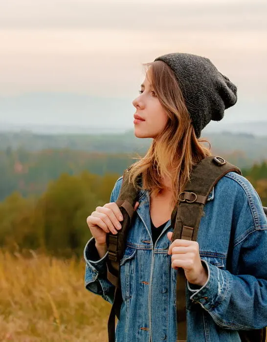 girl-in-denim-jacket-and-hat-with-backpack-in-countryside-with-mountains-on-background