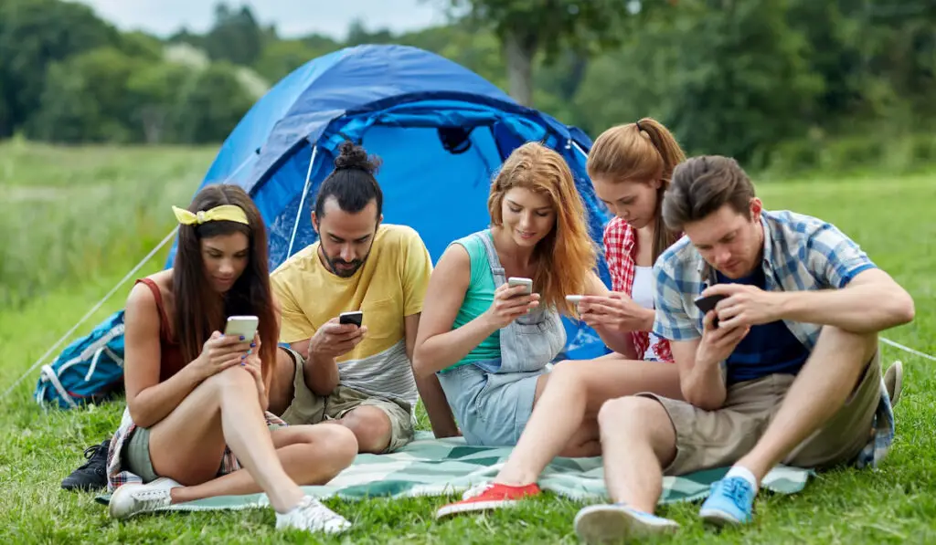 technology and people concept, group of friends with smartphone and tent at camping 