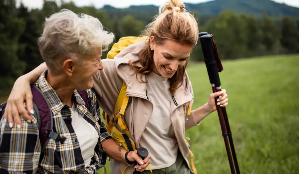 Happy mid adult woman with trekking poles hiking with active senior mother outdoors in nature
