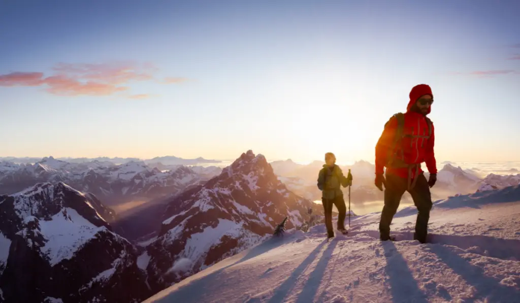 Fantasy Adventure Composite Image of Man and Woman Mountaineering up Snow with Mountain Peaks in Background. Dramatic Sunset or Sunrise. Landscape from British Columbia, Canada.